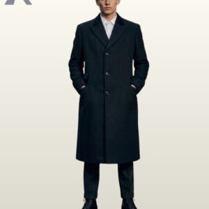 Full front view of a model wearing a buttoned men's black wool long overcoat featuring a black velvet collar accent, peak lapels, and newsboy cap.