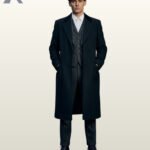 Full front view of a model wearing an open black wool single-breasted overcoat over a grey tweed three-piece suit, pocket watch chain, and newsboy cap.