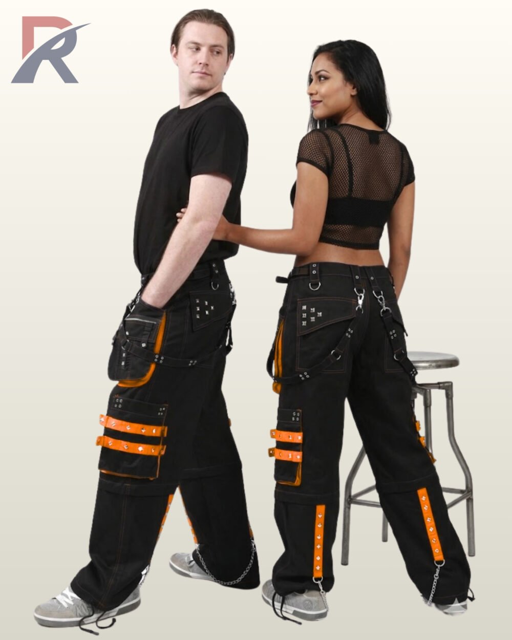 Side and rear view of a couple modeling black gothic cargo trousers with bright orange studded straps, utility pockets, and metal chains.