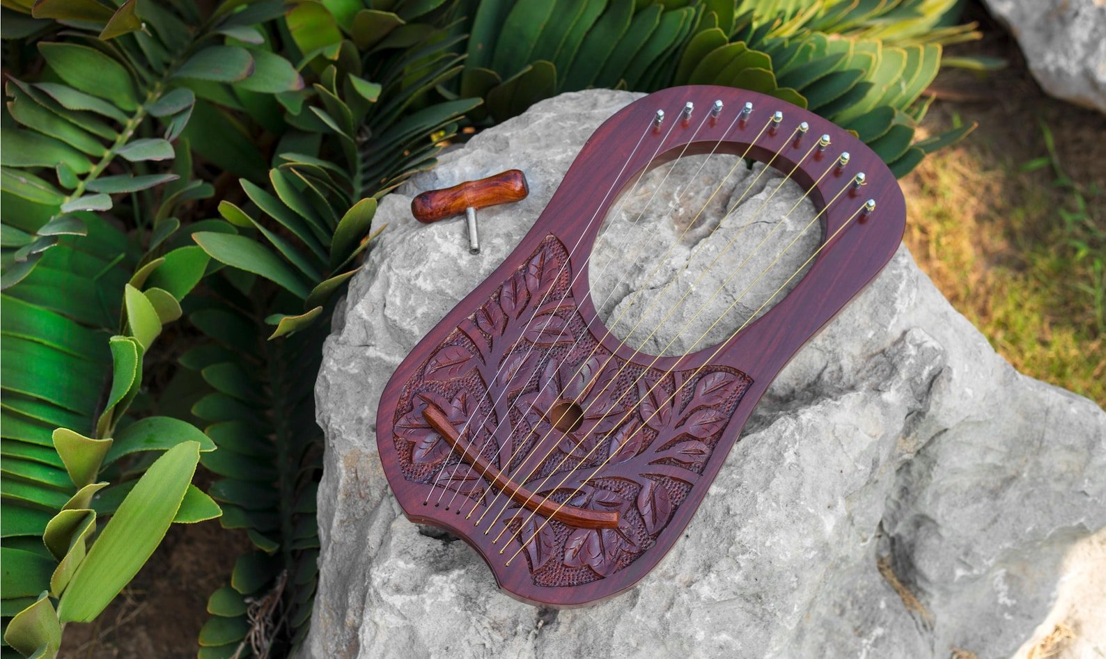 Full front view of a 10-string handcrafted rosewood lyre harp resting on a large stone with a T-shaped wooden tuning key alongside foliage.