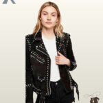 Front view of a model wearing a women's black suede moto biker jacket with silver dome stud embellishments, asymmetrical zip, and waist belt.